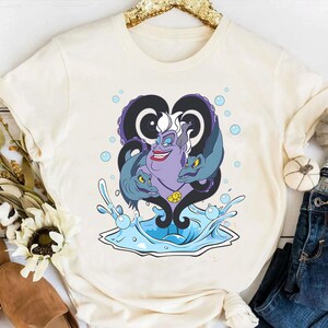 May include: Cream-colored t-shirt featuring a graphic of Ursula from The Little Mermaid. The design includes Ursula's face, tentacles, and eels, with water splashes and bubbles. The artwork is in shades of purple, blue, and black.