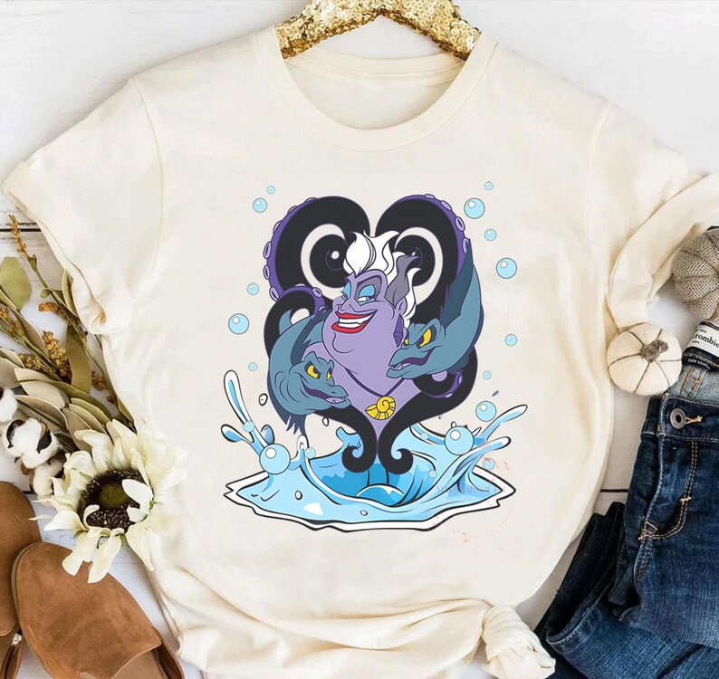 May include: Cream-colored t-shirt featuring a graphic of Ursula from The Little Mermaid. The design includes Ursula's face, tentacles, and eels, with water splashes and bubbles. The artwork is in shades of purple, blue, and black.