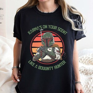 May include: Black t-shirt featuring a graphic of a bounty hunter with a green helmet and armor, set against a sunset background. The text "Karma's on your scent like a bounty hunter" is printed above and below the image.