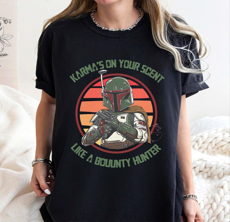 May include: Black t-shirt featuring a graphic of a bounty hunter with a green helmet and armor, set against a sunset background. The text "Karma's on your scent like a bounty hunter" is printed above and below the image.