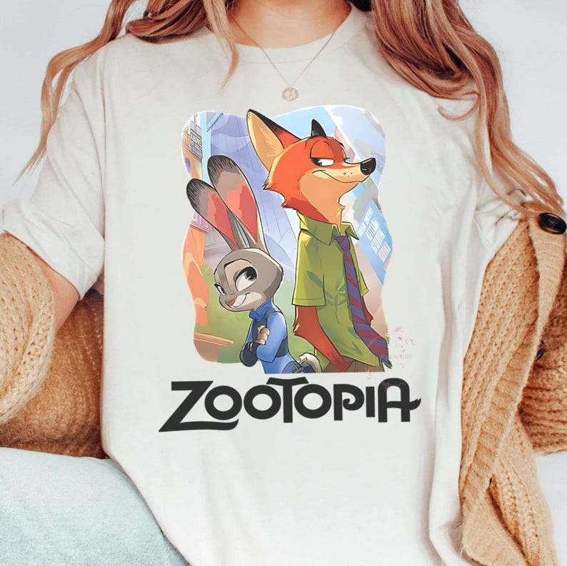 May include: Cream-colored t-shirt featuring a colorful illustration of characters from the animated movie "Zootopia." The design includes a fox and a rabbit, with the word "Zootopia" in bold, black letters below the characters. The shirt has a round neckline.