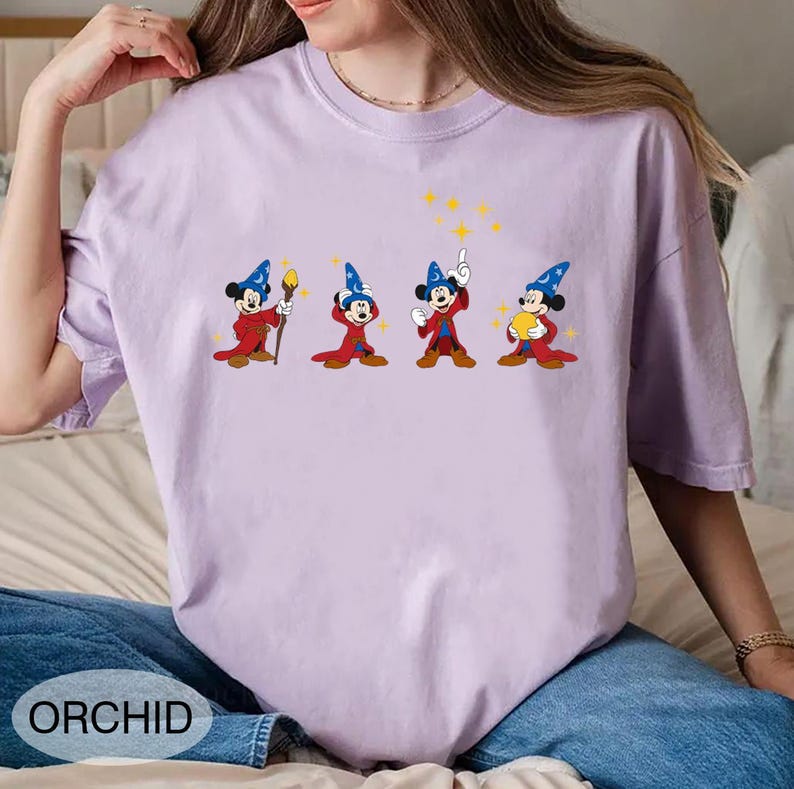 May include: Orchid-colored t-shirt featuring a cartoon graphic of Mickey Mouse in a wizard costume. The graphic shows Mickey in four different poses, with a staff, pointing, and holding a star. The word "ORCHID" is printed on the bottom left.
