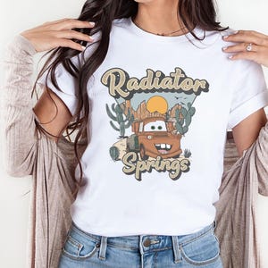 May include: White t-shirt with a graphic of Mater from Cars. The graphic features Mater driving through a desert landscape with the text "Radiator Springs" in a vintage font.