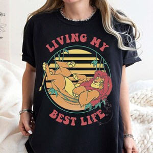 May include: Black t-shirt featuring a cartoon lion lounging in a circle with bamboo accents and the text "LIVING MY BEST LIFE" in red and orange. The lion is orange and yellow.