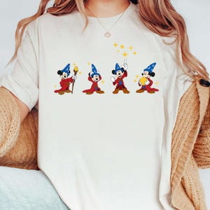 May include: Off-white t-shirt featuring a cartoon graphic of Mickey Mouse in a wizard costume. Mickey is depicted in four different poses, wearing a blue wizard hat and red robe, with yellow star accents.