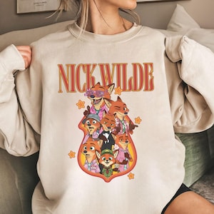 May include: Beige crewneck sweatshirt featuring a graphic of cartoon foxes, with the text "NICK WILDE" in orange and red. The graphic includes multiple fox characters in various outfits and poses, surrounded by orange stars.