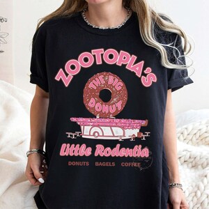 Puede incluir: Camiseta negra con un dise&ntilde;o rosa. El dise&ntilde;o incluye el texto "Zootopia's" y una rosquilla que dice "The Big Donut". Debajo hay un carrito rosa con "Little Rodentia" y las palabras "Donuts Bagels Coffee".
