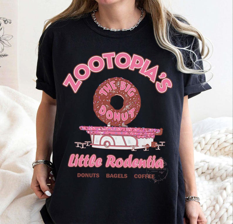 Puede incluir: Camiseta negra con un dise&ntilde;o rosa. El dise&ntilde;o incluye el texto "Zootopia's" y una rosquilla que dice "The Big Donut". Debajo hay un carrito rosa con "Little Rodentia" y las palabras "Donuts Bagels Coffee".