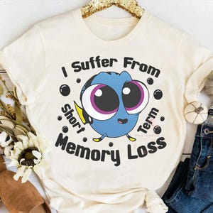 May include: Cream-colored t-shirt featuring a cartoon blue fish with large eyes and the text "I Suffer From Short Term Memory Loss." The design includes black bubbles and yellow accents. The shirt is likely made of a soft, comfortable material.