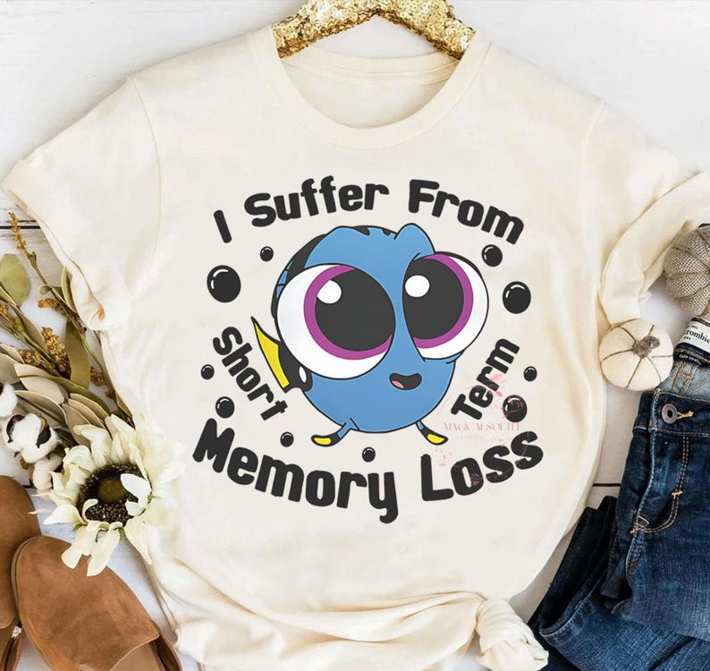 May include: Cream-colored t-shirt featuring a cartoon blue fish with large eyes and the text "I Suffer From Short Term Memory Loss." The design includes black bubbles and yellow accents. The shirt is likely made of a soft, comfortable material.