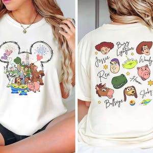 May include: Cream-coloured t-shirt featuring a Mickey Mouse head outline with fireworks and Toy Story characters. The back of the shirt displays character faces and names like Woody, Buzz Lightyear, and Jessie.