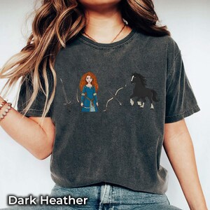 May include: Dark heather gray t-shirt featuring a cartoon illustration of a red-haired woman in a blue dress, a black horse, a sword, and a bow and arrow. The text "Dark Heather" is at the bottom.