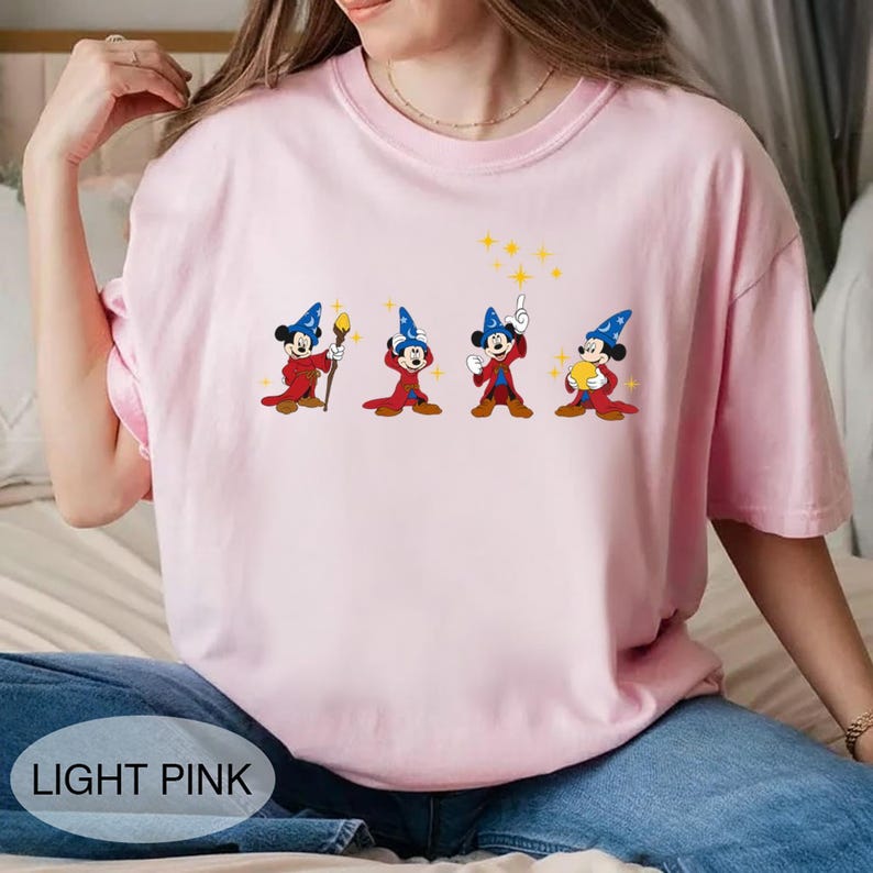 May include: Light pink t-shirt featuring a graphic of Mickey Mouse in a wizard costume. The design shows Mickey in four different poses with a blue wizard hat, red robe, and yellow accents. The text "LIGHT PINK" is at the bottom.