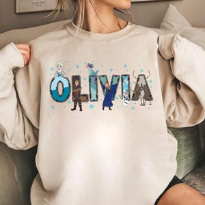 Pu&ograve; includere: Felpa girocollo beige con il nome "OLIVIA" in lettere blu e turchese, decorata con personaggi dei cartoni animati e fiocchi di neve. Il design &egrave; ispirato a un popolare film d'animazione.