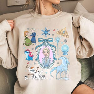 May include: Beige sweatshirt with cartoon illustrations of characters and symbols from a popular animated film. The design includes a princess, a snowman, a horse, and snowflakes. The illustrations are in shades of blue, white, and pink.