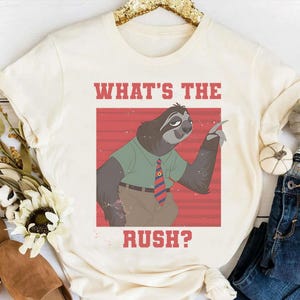 May include: Cream-colored t-shirt featuring a cartoon sloth wearing a green shirt and a red and blue striped tie. The text "WHAT'S THE RUSH?" is printed in red above and below the sloth illustration. The shirt is casual wear.