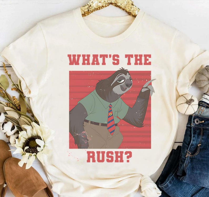 May include: Cream-colored t-shirt featuring a cartoon sloth wearing a green shirt and a red and blue striped tie. The text "WHAT'S THE RUSH?" is printed in red above and below the sloth illustration. The shirt is casual wear.