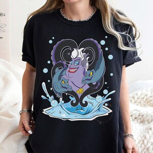 May include: Black t-shirt featuring a cartoon illustration of Ursula from The Little Mermaid. The purple character has white hair, a red smile, and is surrounded by two eel-like creatures. Blue water and bubbles complete the design.