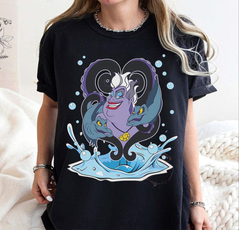 May include: Black t-shirt featuring a cartoon illustration of Ursula from The Little Mermaid. The purple character has white hair, a red smile, and is surrounded by two eel-like creatures. Blue water and bubbles complete the design.
