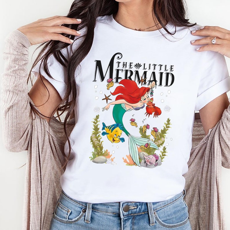Op de afbeelding: Wit T-shirt met een afbeelding van Ariel, de Kleine Zeemeermin, uit de Disney-film. De afbeelding toont Ariel die zwemt met Vlotter en Sebastian. De tekst "The Little Mermaid" is in zwart boven de afbeelding gedrukt.