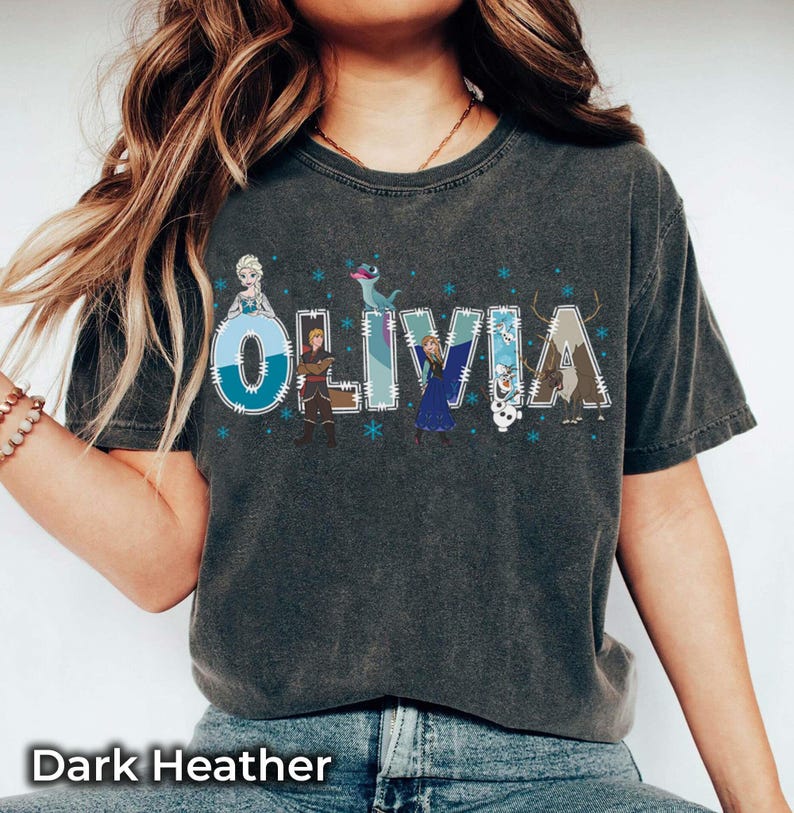 Pu&ograve; includere: T-shirt grigio scuro con il nome "OLIVIA" in un design stravagante con personaggi dei cartoni animati e fiocchi di neve. Le lettere sono decorate con immagini di personaggi dei cartoni animati. Il testo "Dark Heather" &egrave; in basso.