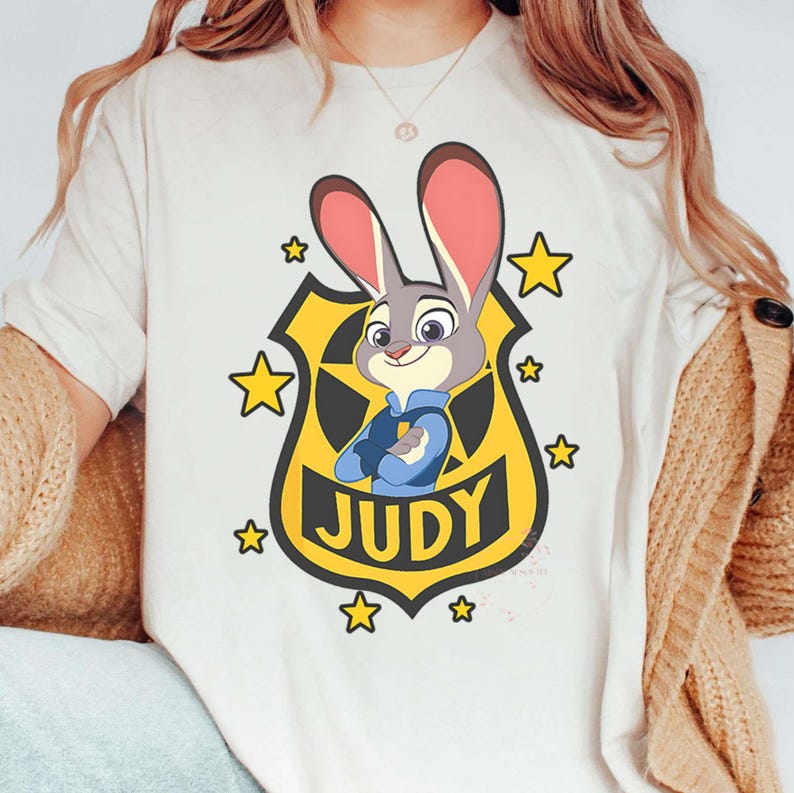 May include: Cream-colored t-shirt featuring a cartoon image of Judy Hopps from Zootopia. Judy is depicted in a police badge with the name "JUDY" below. The design includes yellow stars and a yellow badge.