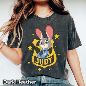 May include: Dark heather gray t-shirt featuring a cartoon image of Judy Hopps from Zootopia. The design includes Judy in a police badge, surrounded by yellow stars, with the name "JUDY" below. The text "Dark Heather" is at the bottom.