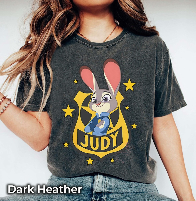 May include: Dark heather gray t-shirt featuring a cartoon image of Judy Hopps from Zootopia. The design includes Judy in a police badge, surrounded by yellow stars, with the name "JUDY" below. The text "Dark Heather" is at the bottom.