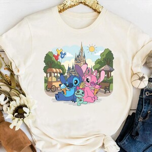 K&ouml;nnte beinhalten: Cremefarbenes T-Shirt mit einem farbenfrohen Aufdruck von Stitch und Angel, die Eis vor einem Schloss genie&szlig;en. Das Design umfasst eine kleine gr&uuml;ne Puppe und bunte Luftballons, vor einem sonnigen Himmel und Park.