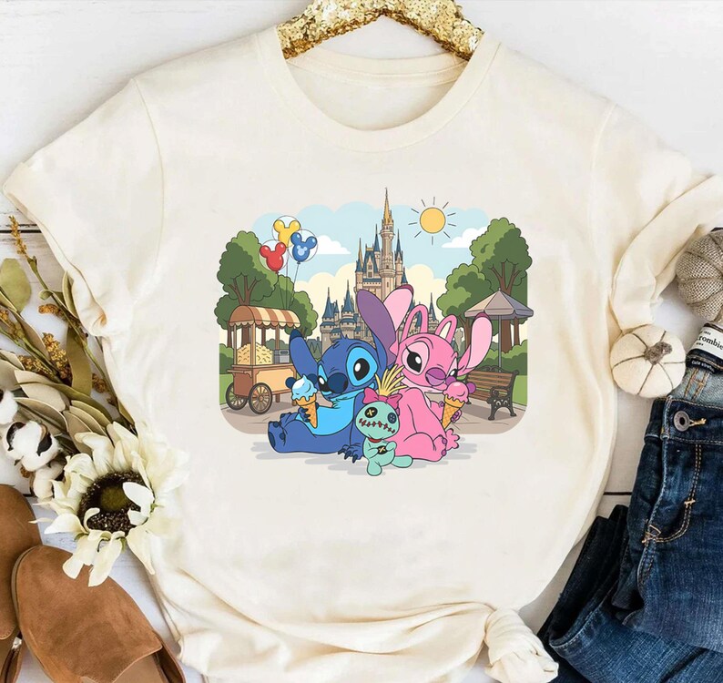 K&ouml;nnte beinhalten: Cremefarbenes T-Shirt mit einem farbenfrohen Aufdruck von Stitch und Angel, die Eis vor einem Schloss genie&szlig;en. Das Design umfasst eine kleine gr&uuml;ne Puppe und bunte Luftballons, vor einem sonnigen Himmel und Park.