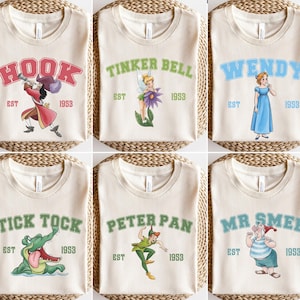 May include: Six cream-colored t-shirts featuring characters from Peter Pan. Each shirt has a different character name in a collegiate font, with an illustration and the text "EST 1953". Characters include Hook, Tinker Bell, Wendy, Tick Tock, Peter Pan, and Mr. Smee.
