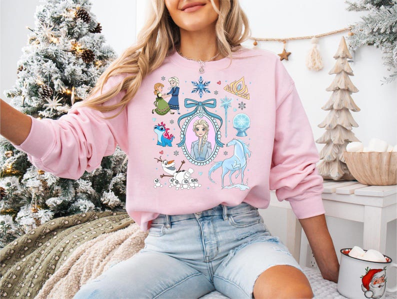 May include: A pale pink sweatshirt with a colourful graphic of animated film characters and symbols. The design includes a portrait, snowflakes, and related imagery. The sweatshirt has long sleeves and a crew neck.