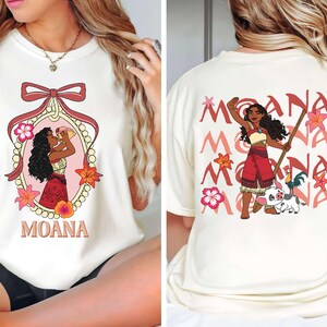 May include: Cream-coloured t-shirt with front and back designs. The front features a cartoon illustration of a woman in a decorative frame with the word "MOANA". The back displays the word "MOANA" repeated with a cartoon character.