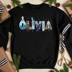 Pu&ograve; includere: Felpa nera con il nome "Olivia" in un carattere decorativo, con personaggi di un popolare film d'animazione. Le lettere sono decorate con fiocchi di neve e illustrazioni di personaggi. La felpa &egrave; circondata da regali confezionati e rami di pino.