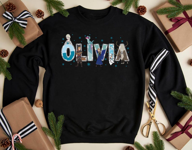Pu&ograve; includere: Felpa nera con il nome "Olivia" in un carattere decorativo, con personaggi di un popolare film d'animazione. Le lettere sono decorate con fiocchi di neve e illustrazioni di personaggi. La felpa &egrave; circondata da regali confezionati e rami di pino.