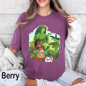 Puede incluir: Camiseta color bayas con un gr&aacute;fico colorido de una mujer con vestimenta tradicional, rodeada de plantas tropicales y un cerdo. El gr&aacute;fico incluye una figura verde y la palabra "Berry" en blanco.