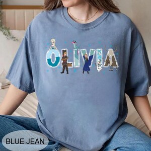 Pu&ograve; includere: T-shirt azzurra con il nome "OLIVIA" in grandi lettere stilizzate, decorata con personaggi dei cartoni animati. La maglietta ha un girocollo e maniche corte. Il testo "BLUE JEAN" &egrave; nella parte inferiore dell'immagine.