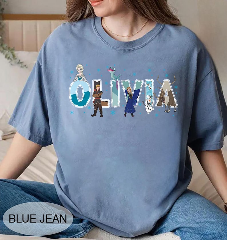 Pu&ograve; includere: T-shirt azzurra con il nome "OLIVIA" in grandi lettere stilizzate, decorata con personaggi dei cartoni animati. La maglietta ha un girocollo e maniche corte. Il testo "BLUE JEAN" &egrave; nella parte inferiore dell'immagine.