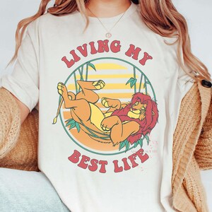 May include: Cream-colored t-shirt featuring a cartoon lion resting inside a circular design with bamboo stalks. The text "LIVING MY BEST LIFE" is arched above and below the lion illustration. The lion is orange and yellow with a red mane.