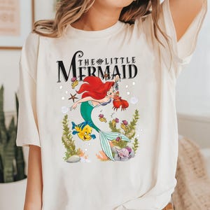 Op de afbeelding: Wit T-shirt met een afbeelding van Ariel, de Kleine Zeemeermin, die zwemt met Vlotter en Sebastian. De tekst "De Kleine Zeemeermin" staat boven de afbeelding.