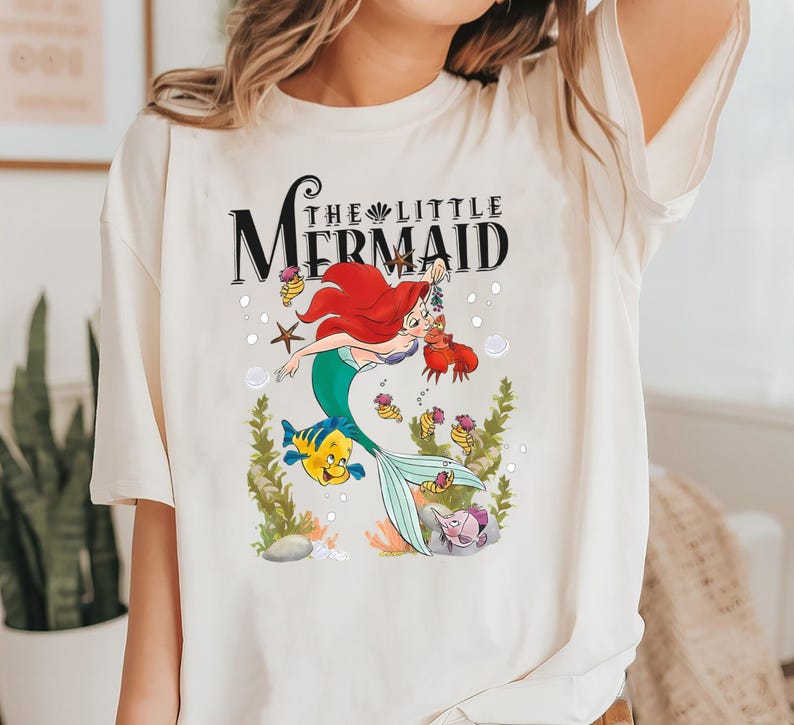 Op de afbeelding: Wit T-shirt met een afbeelding van Ariel, de Kleine Zeemeermin, die zwemt met Vlotter en Sebastian. De tekst "De Kleine Zeemeermin" staat boven de afbeelding.