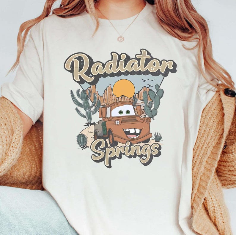 May include: A white t-shirt with a vintage-style graphic of a rusty brown truck with a smiling face. The graphic says "Radiator Springs" with a sunset and cacti in the background.