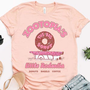 Puede incluir: Camiseta rosa claro con un dise&ntilde;o gr&aacute;fico. El dise&ntilde;o incluye el texto "Zootopia's The Big Donut" sobre una ilustraci&oacute;n de donut. Debajo, el texto "Little Rodentia" y las palabras "Donuts Bagels Coffee".