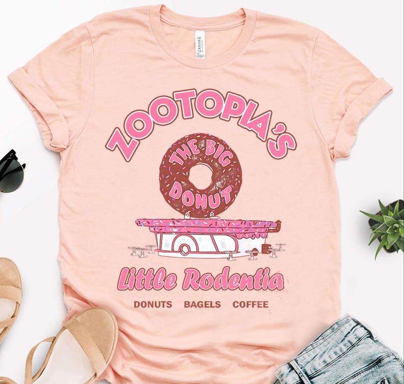 Puede incluir: Camiseta rosa claro con un dise&ntilde;o gr&aacute;fico. El dise&ntilde;o incluye el texto "Zootopia's The Big Donut" sobre una ilustraci&oacute;n de donut. Debajo, el texto "Little Rodentia" y las palabras "Donuts Bagels Coffee".