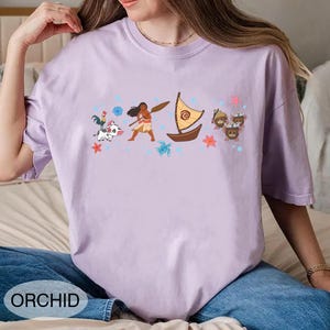 May include: A lilac t-shirt with a cartoon graphic featuring a woman with a paddle, a boat, and other characters. The word "ORCHID" is printed at the bottom. The shirt is made of a soft material.
