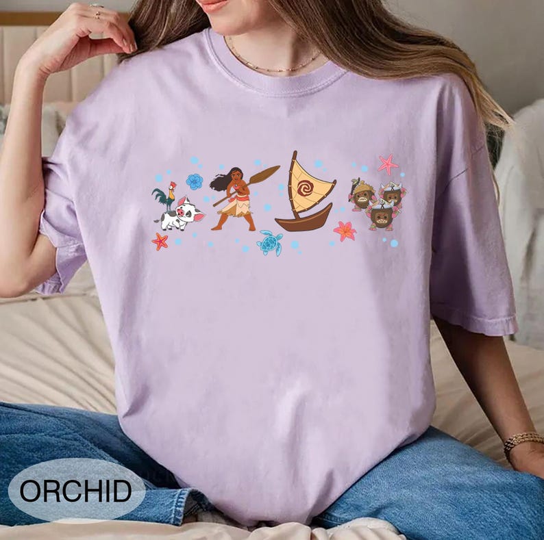 May include: A lilac t-shirt with a cartoon graphic featuring a woman with a paddle, a boat, and other characters. The word "ORCHID" is printed at the bottom. The shirt is made of a soft material.