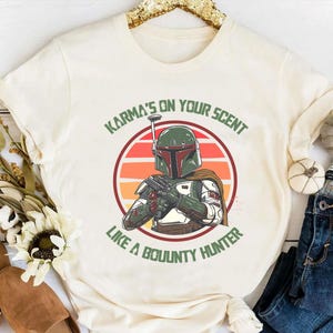 May include: Cream-colored t-shirt featuring a graphic of a bounty hunter in a green helmet and armor, set against a sunset background. The text "Karma's on your scent like a bounty hunter" is printed above and below the image.