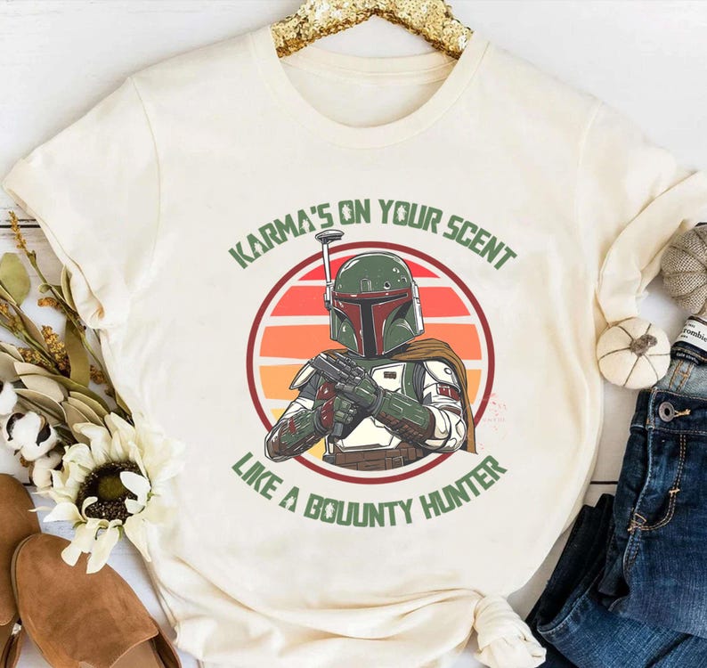 May include: Cream-colored t-shirt featuring a graphic of a bounty hunter in a green helmet and armor, set against a sunset background. The text "Karma's on your scent like a bounty hunter" is printed above and below the image.