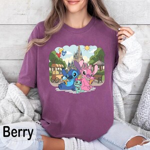 K&ouml;nnte beinhalten: Beerenfarbenes T-Shirt mit einer Grafik von Stitch, Angel und einem kleinen gr&uuml;nen Wesen, das Eis vor einem Schloss genie&szlig;t. Das Design umfasst einen Essenswagen, Luftballons und einen sonnigen Himmel. Das Wort "Berry" ist unten aufgedruckt.