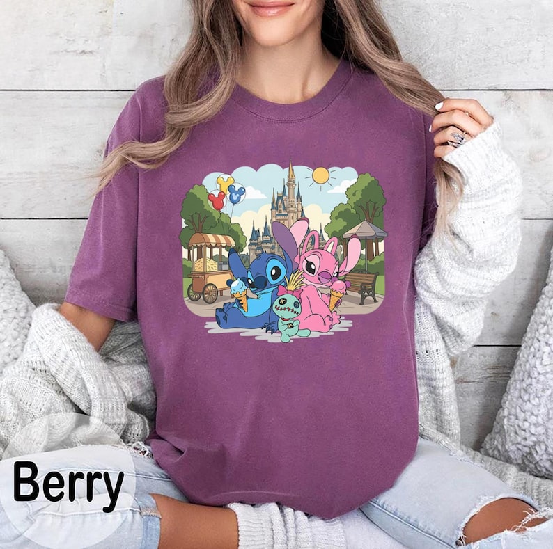 K&ouml;nnte beinhalten: Beerenfarbenes T-Shirt mit einer Grafik von Stitch, Angel und einem kleinen gr&uuml;nen Wesen, das Eis vor einem Schloss genie&szlig;t. Das Design umfasst einen Essenswagen, Luftballons und einen sonnigen Himmel. Das Wort "Berry" ist unten aufgedruckt.